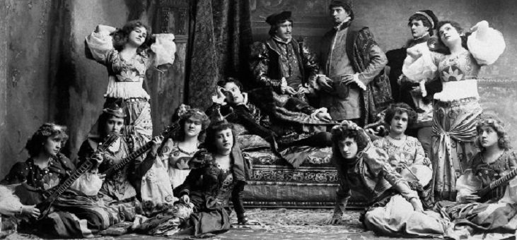 Two men at the back balance on a couch. Two standing ladies are to the right of them, one to the left, and a number are sitting on the floor in front of them.