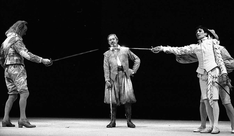 Aguecheek and "Sebastian" (Viola) point swords at each other, far enough apart that even the fully extended swords do not touch. Viola is being encouraged by someone standing behind her, while in the rear stands a man with a drawn sword held pointing at the ground.