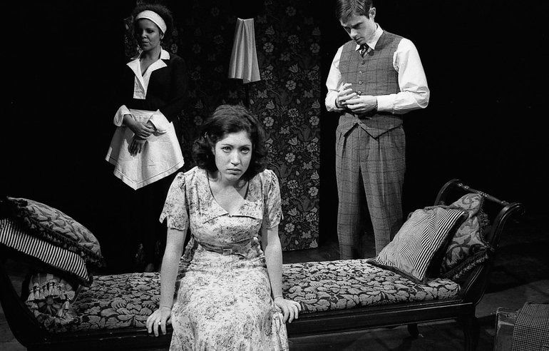 Julia in a modern dress sits on a chaise longue looking unhappy. Behind to her left Proteus in a suit (but not wearing a jacket) is twisting a ring on his finger. Behind Julia on the other side stands her maid, dressed in black with a white apron.