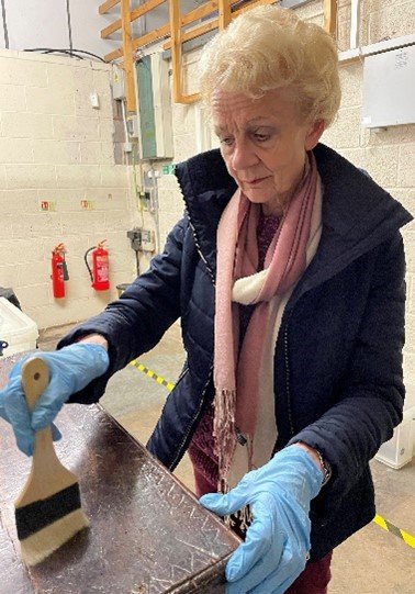 Gail dusting a wooden dark wood chest from the 1600s, the brush she is using looks like a large flat paintbrush with white bristles.