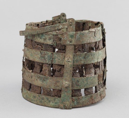An Anglo-Saxon situla which looks recognisably like a bucket, with decayed wooden sides bound with corroded metal strips. There is a hinged handle at the top.
