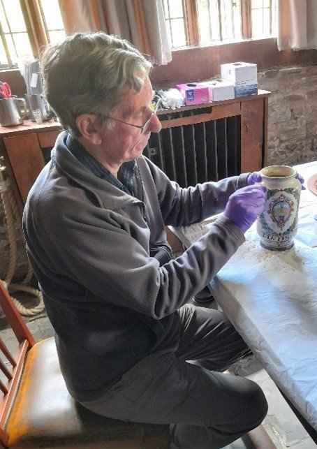 Volunteer Rodger cleaning a medicine jar from the 1600s. He is sat at a large table and is wearing purple gloves. The medicine jar is white with green decoration.